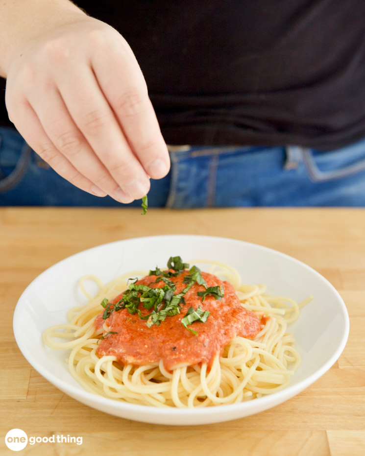 pasta sauce hacks Photo showing a hand sprinkling chopped basil on a plate of spaghetti with pasta sauce.